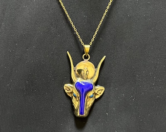 May include: Gold-coloured necklace with a pendant depicting a bull-like figure. The pendant incorporates blue enamel and a sun-shaped element. The necklace is set against a dark backdrop, emphasising the jewellery's intricate design.
