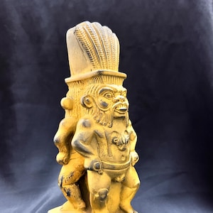 Może przedstawiać: Szczegółowa, złota figurka bóstwa z pióropuszem i surowym wyrazem twarzy. Postać ma wiele ramion i stoi na prostokątnej podstawie z grawerowanymi detalami. Posąg jest prawdopodobnie przedmiotem dekoracyjnym.