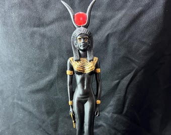 May include: A black statue of an Egyptian deity, with gold detailing on the necklace, armbands, and feet. The figure wears a headdress with a red orb and horns. The statue stands on a rectangular base with gold hieroglyphs.