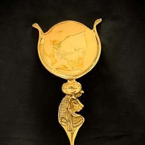 May include: A gold-colored decorative item with an Egyptian theme. The item features a circular top with curved handles, a detailed lion's head, and a long, slender handle. The design is intricate and appears to be made of metal.