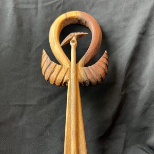 May include: A hand-carved wooden sculpture featuring a stylized bird with outstretched wings. The sculpture is made from various shades of brown wood, with a circular element above the bird's head. The wings have carved details, and the overall design is symmetrical.