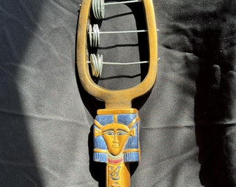May include: A gold-colored wooden musical instrument with a carved Egyptian goddess head and hieroglyphics. The instrument has a frame with four metal rings that make a rattling sound when shaken.