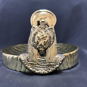 May include: A gold-colored bracelet with an Egyptian-inspired design. The centerpiece features a lion's head with a sun disc above it. The band has a textured, patterned design.