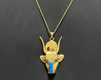 May include: Gold-coloured pendant necklace featuring a bull's head with blue accents. The pendant hangs from a delicate chain. The bull's head has detailed horns and ears, and a blue stripe down the centre.