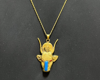 May include: A gold-coloured pendant necklace with a bull's head design. The pendant has a blue inlay and red accents. The necklace chain is delicate and gold, with a lobster clasp. The necklace is displayed against a dark background.
