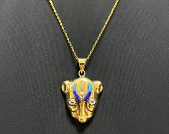 Puede incluir: Un collar con colgante dorado con un diseño de cabeza de animal estilizada, que incorpora detalles azules y blancos. El colgante cuelga de una fina cadena dorada. El collar se muestra sobre un fondo negro.