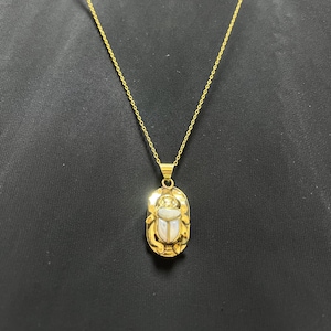 May include: Gold-toned necklace featuring a pendant with an oval frame and a scarab beetle design. The beetle is white and the frame is gold. The necklace has a delicate chain and a clasp. The background is black.