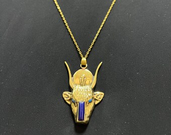 May include: Gold-coloured pendant necklace featuring a stylised bull's head with blue and gold accents. The pendant has a sun disc above the head and a blue vertical stripe. The necklace chain is delicate and gold-coloured.