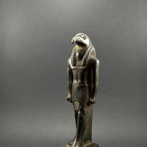 May include: Black statue of the Egyptian god Horus, depicted as a man with a falcon head. The statue is standing on a rectangular base with hieroglyphs.