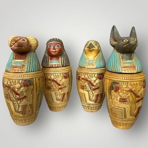 Puede incluir: Cuatro jarras canopias egipcias decorativas. Cada jarra presenta una cabeza de animal única, incluyendo un babuino, un humano, un halcón y un chacal. Las jarras están pintadas en oro, rojo y turquesa, con tallas detalladas de figuras y jeroglíficos. Estas jarras son una representación del arte egipcio antiguo.