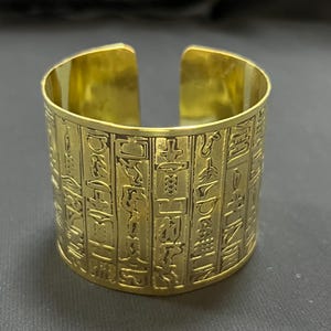 Könnte beinhalten: Ein goldfarbenes Armband mit eingravierten Hieroglyphen. Das offene Armband hat eine zylindrische Form und besteht aus glänzendem Metall. Die Hieroglyphen sind vertikal um das Armband angeordnet.