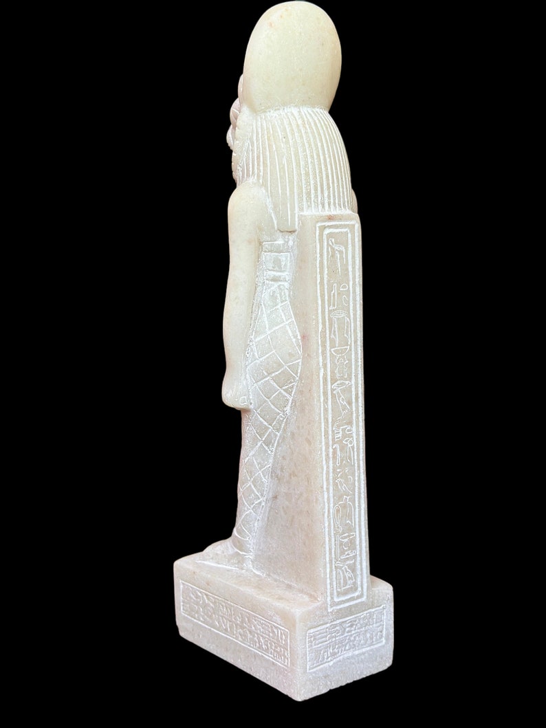 May include: White alabaster statue of an Egyptian god with hieroglyphics on the base and side. The statue is standing on a rectangular base.