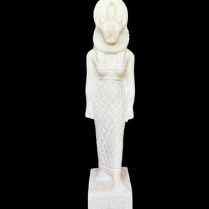 May include: White stone statue of a female figure with a lion head and a sun disk on her head. The statue is standing on a rectangular base with hieroglyphs.