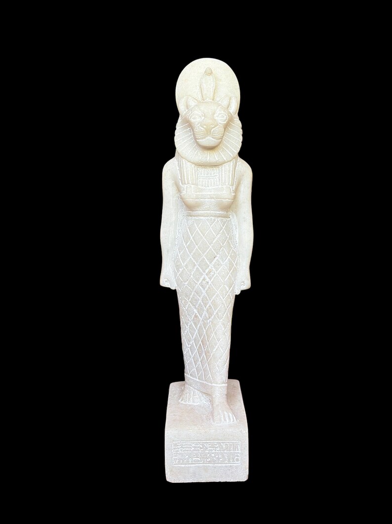 May include: White stone statue of a female figure with a lion head and a sun disk on her head. The statue is standing on a rectangular base with hieroglyphs.