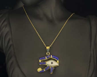 Puede incluir: Collar dorado con un colgante con forma de Ojo de Horus. El colgante tiene un borde esmaltado azul, un ojo blanco y detalles dorados. El collar se muestra sobre un maniquí negro.