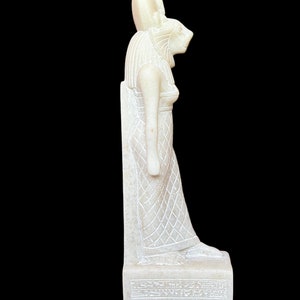 May include: White alabaster statue of the Egyptian goddess Bastet, depicted as a woman with a lion's head, standing on a base with hieroglyphs.