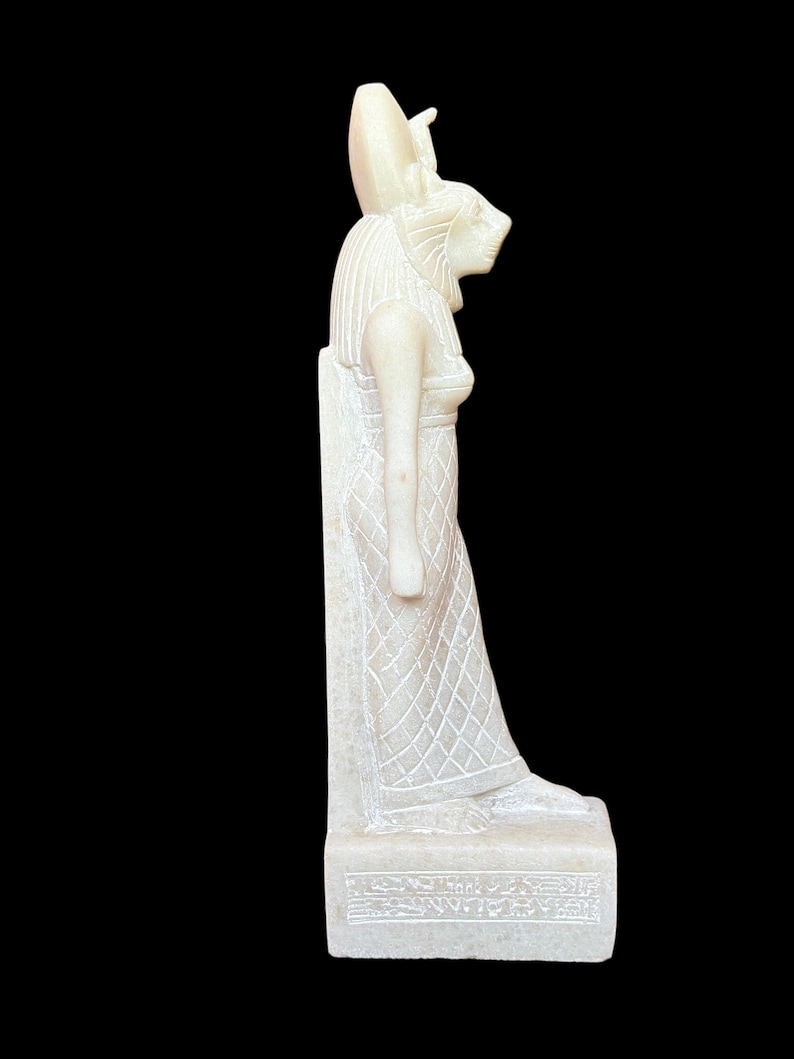May include: White alabaster statue of the Egyptian goddess Bastet, depicted as a woman with a lion's head, standing on a base with hieroglyphs.