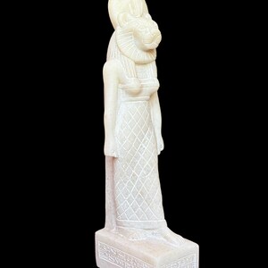 May include: White alabaster statue of the Egyptian goddess Sekhmet, depicted as a lion-headed woman wearing a crown and a pleated dress. The statue stands on a rectangular base with hieroglyphic inscriptions.