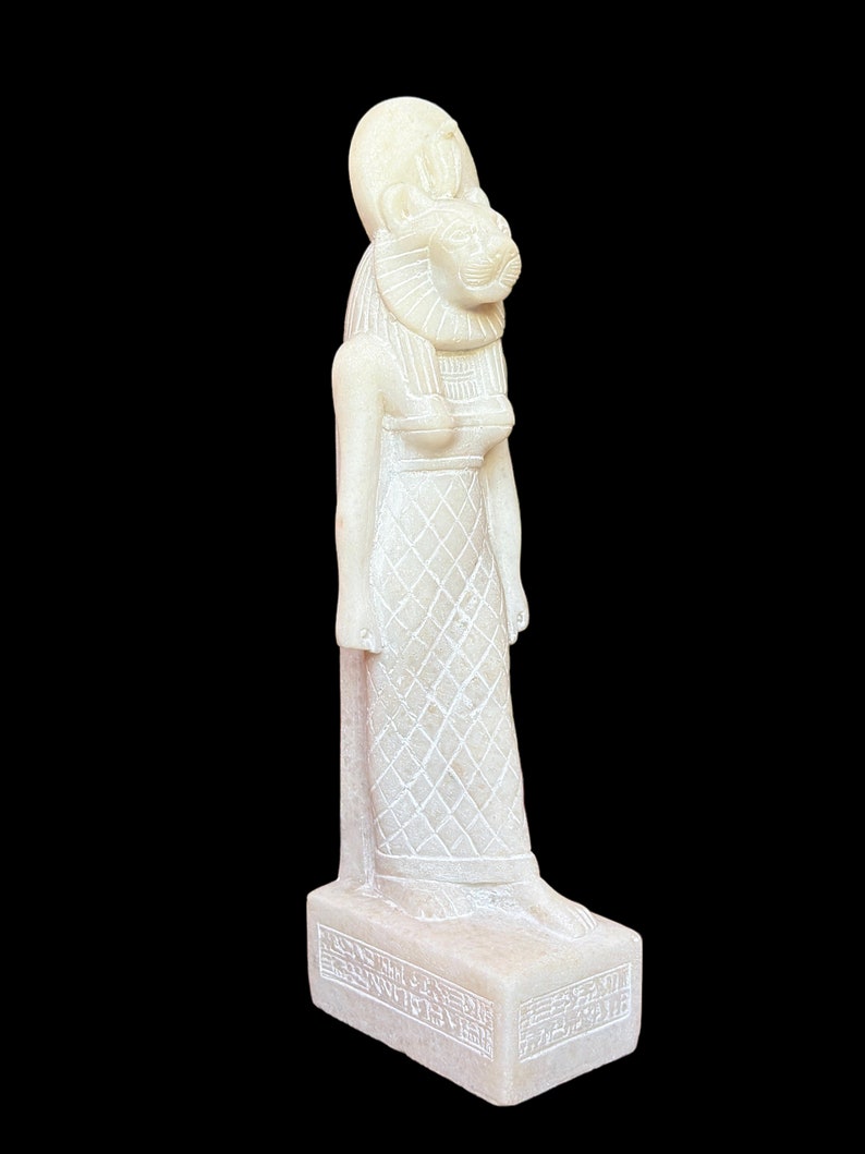 May include: White alabaster statue of the Egyptian goddess Sekhmet, depicted as a lion-headed woman wearing a crown and a pleated dress. The statue stands on a rectangular base with hieroglyphic inscriptions.