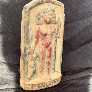 May include: A terracotta relief sculpture of a female figure with red and black paint. The figure is standing with her arms raised, holding a staff in one hand and a bird in the other. The sculpture is framed by a rectangular border.