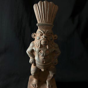 Op de afbeelding: Een bronzen beeldje van een hurkende figuur met een gedetailleerde hoofdtooi. De figuur heeft een strenge uitdrukking, armen over elkaar, en staat op een rechthoekige basis met geëtste ontwerpen. Het beeld heeft een gedetailleerd gezicht en lichaam.