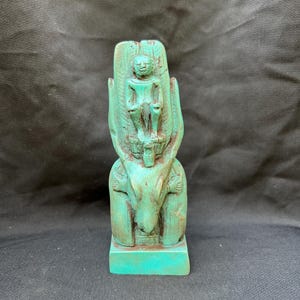 Puede incluir: Una escultura tallada de color turquesa de una figura sentada sobre un animal o criatura estilizada. La escultura tiene detalles intrincados y parece estar hecha de un material sólido. El fondo es una tela oscura y texturizada.