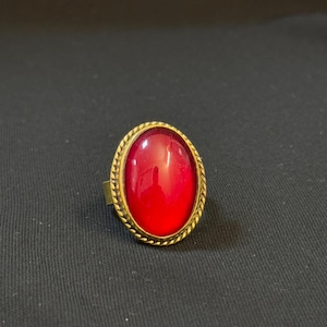 Może przedstawiać: Złoty pierścionek z dużym, owalnym, czerwonym kamieniem szlachetnym.
