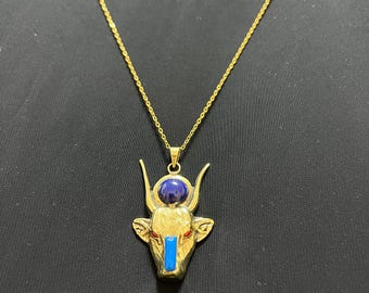 May include: Gold-coloured necklace with a pendant depicting a bull's head. The pendant incorporates a blue stone and a blue vertical stripe. The necklace is set against a black backdrop.