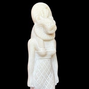 May include: White alabaster statue of an Egyptian goddess with a lion head and a crown. The goddess is wearing a long, flowing dress with a geometric pattern.