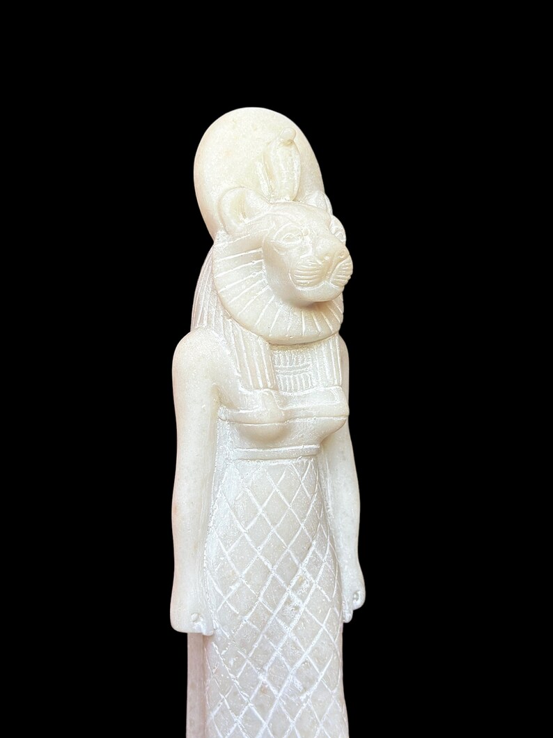 May include: White alabaster statue of an Egyptian goddess with a lion head and a crown. The goddess is wearing a long, flowing dress with a geometric pattern.
