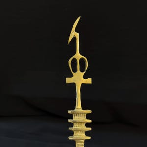 Può includere: Una scultura in metallo dorato di un ankh, un simbolo della vita nell'antico Egitto, con una punta appuntita e una base a più livelli.
