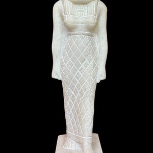 May include: White alabaster statue of the Egyptian goddess Bastet, depicted as a woman with a lion's head. She is wearing a traditional Egyptian dress with a patterned skirt and a crown. The statue is standing on a rectangular base.
