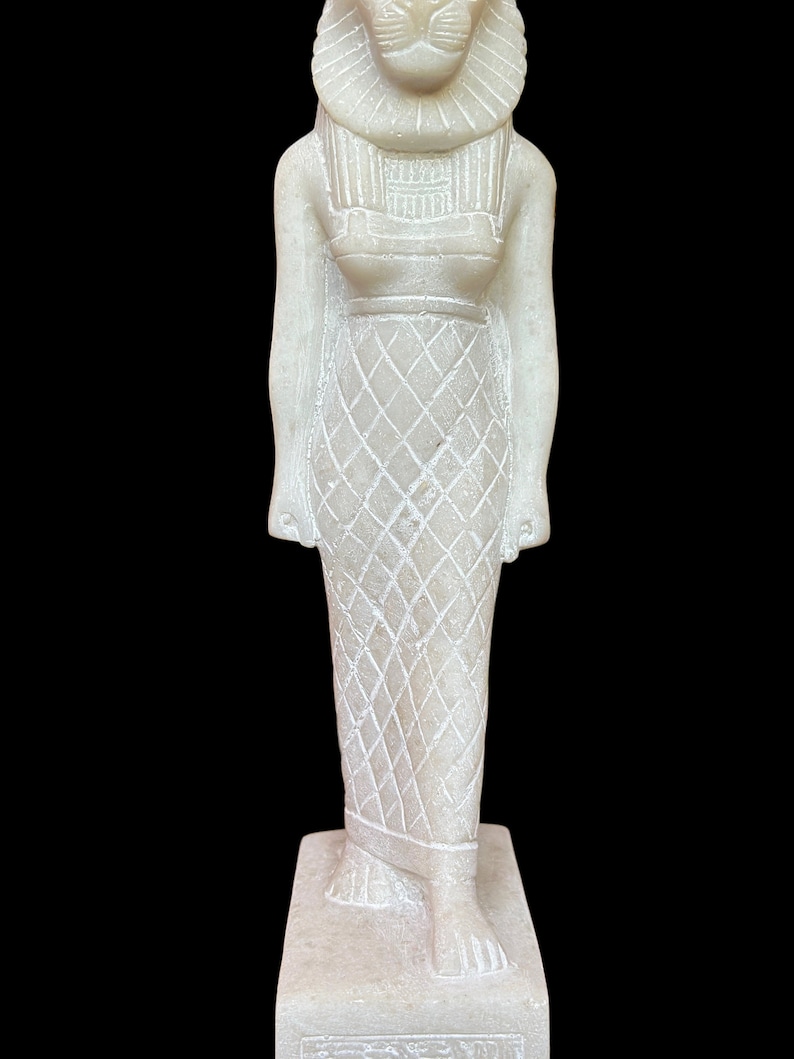 May include: White alabaster statue of the Egyptian goddess Bastet, depicted as a woman with a lion's head. She is wearing a traditional Egyptian dress with a patterned skirt and a crown. The statue is standing on a rectangular base.