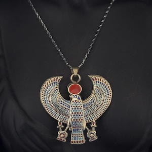 May include: A gold-toned necklace featuring a detailed pendant depicting a winged falcon with red, blue, and gold enamel accents. The pendant is suspended from a silver chain.