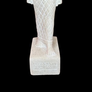 May include: White marble statue of a standing figure wearing a patterned garment. The statue is on a rectangular base with hieroglyphics inscribed on the front.