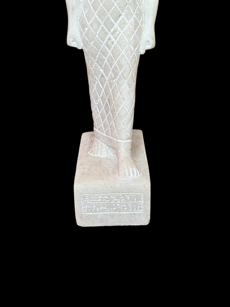 May include: White marble statue of a standing figure wearing a patterned garment. The statue is on a rectangular base with hieroglyphics inscribed on the front.