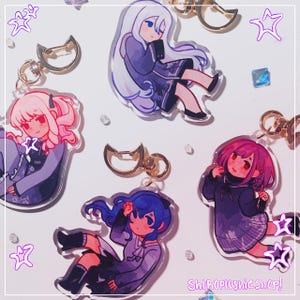 Może przedstawiać: Akrylowe breloczki do kluczy z postaciami w stylu anime w różnych pozach. Każdy breloczek ma złote zapięcie. Postacie mają różne kolory włosów, w tym różowy, niebieski i biały, i noszą ciemne stroje. Widoczny jest tekst "ShiroPlushie shop!".