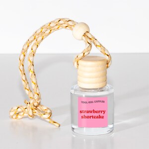 Peut inclure: Une bouteille en verre transparente avec un bouchon en bois et une ficelle pour l'accrocher. La bouteille contient un d&eacute;sodorisant pour voiture avec le texte "Cool Girl Candles" et "strawberry shortcake" sur l'&eacute;tiquette.