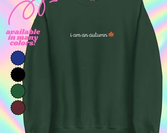 Könnte beinhalten: Ein dunkelgrünes Sweatshirt mit dem Schriftzug "i am an autumn" in weißer Schrift, begleitet von einer orangefarbenen Blattgrafik. Das Bild zeigt auch eine Farbpalette aus Blau, Schwarz, Grün und Burgunderrot, mit dem Text "available in many colors!"