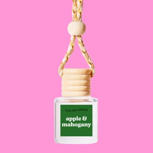 May include: A clear glass bottle with a green label that says "cool car candles" and "apple & mahogany". The bottle is hanging from a brown and gold rope.