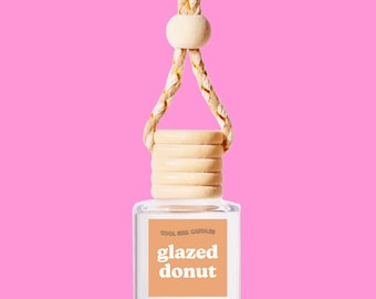 May include: A clear glass bottle with a wooden cap and a string for hanging. The bottle contains a car air freshener with the text "Cool Girl Candles glazed donut" on the label.