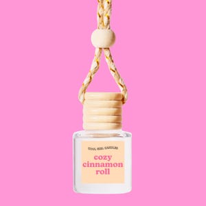 May include: A clear, square-shaped car air freshener with a wooden cap and a braided rope hanger. The label reads "cozy cinnamon roll" in pink text on a peach background, with "COOL GIRL CANDLES" above. The background is a solid pink.