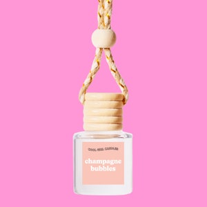 May include: A clear glass bottle with a wooden cap and a gold string hanging from it. The bottle has a label that says "Cool Girl Candles" and "champagne bubbles".