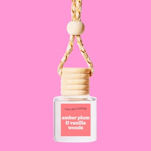 May include: A clear glass bottle with a wooden cap and a gold string hanging from it. The bottle has a label that says "Cool Girl Candles" and "amber plum & vanilla woods".