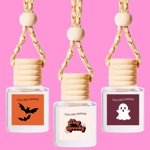 May include: Three glass car air fresheners with a wooden top and a string for hanging. The air fresheners are decorated with Halloween themes. The first one has an orange background with two black bats. The second one has a white background with the text "Stay Spooky" in orange and black. The third one has a maroon background with a white ghost.