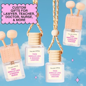 Puede incluir: Cuatro ambientadores para coche de cristal con tapas de madera y cordones para colgar. Cada botella tiene una etiqueta que dice "This Smells Like a Badass" seguida de "Lawyer", "Nurse", "Doctor" o "Teacher". Un gráfico de burbujas rosa dice "Custom Gifts for Lawyer, Teacher, Doctor, Nurse, & More".