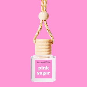 Puede incluir: Una botella de vidrio transparente con una tapa de madera y una cuerda para colgar. La botella contiene un ambientador de coche con aroma a azúcar rosa. La etiqueta dice "Cool Girl Candles" y "pink sugar".
