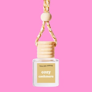 Puede incluir: Un ambientador para coche de cristal transparente con tapón de madera y cuerda trenzada. La etiqueta muestra "COZY CASHMERE" en una fuente suave. El fondo es de color rosa brillante.