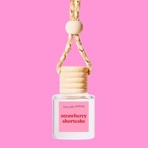 Puede incluir: Una botella de vidrio transparente con una tapa de madera y una cuerda para colgar. La botella tiene una etiqueta que dice "Cool Girl Candles" y "strawberry shortcake".
