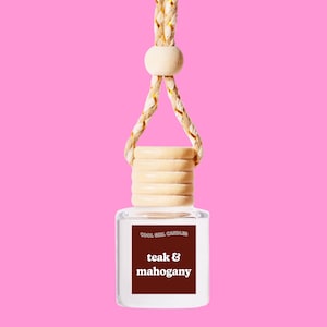 May include: A clear glass bottle with a wooden cap and a string for hanging. The bottle contains a car air freshener with the text "COOL GIRL CANDLES teak & mahogany".
